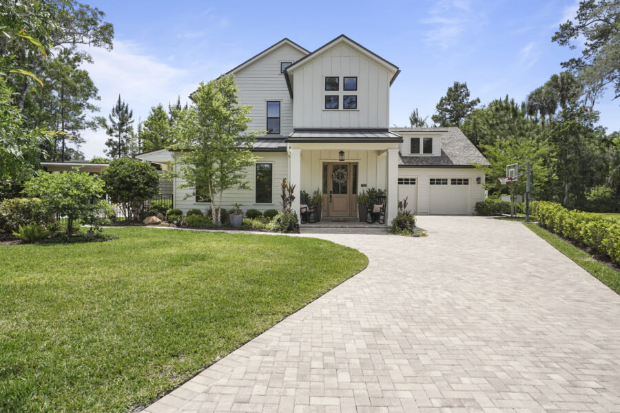 Modern white farmhouse-style home with a landscaped front yard, paved driveway, and a basketball hoop, surrounded by lush trees and greenery under a clear blue sky.
