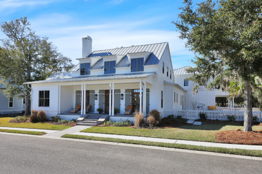 Modern white farmhouse-style home with a metal roof, front porch, and lush landscaping in a suburban neighborhood.