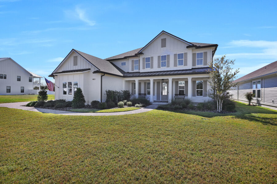 Spacious white two-story farmhouse-style home with front porch, surrounded by a well-maintained lawn and landscaping, under a clear blue sky.