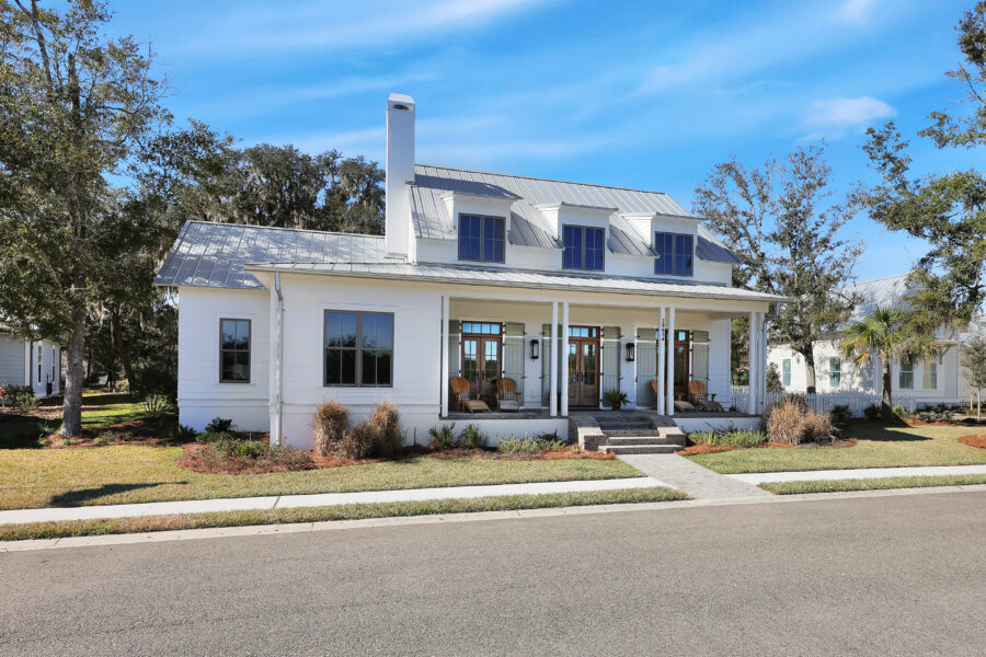 Modern white farmhouse with a metal roof, front porch with seating, surrounded by landscaped yard and trees under a clear blue sky.