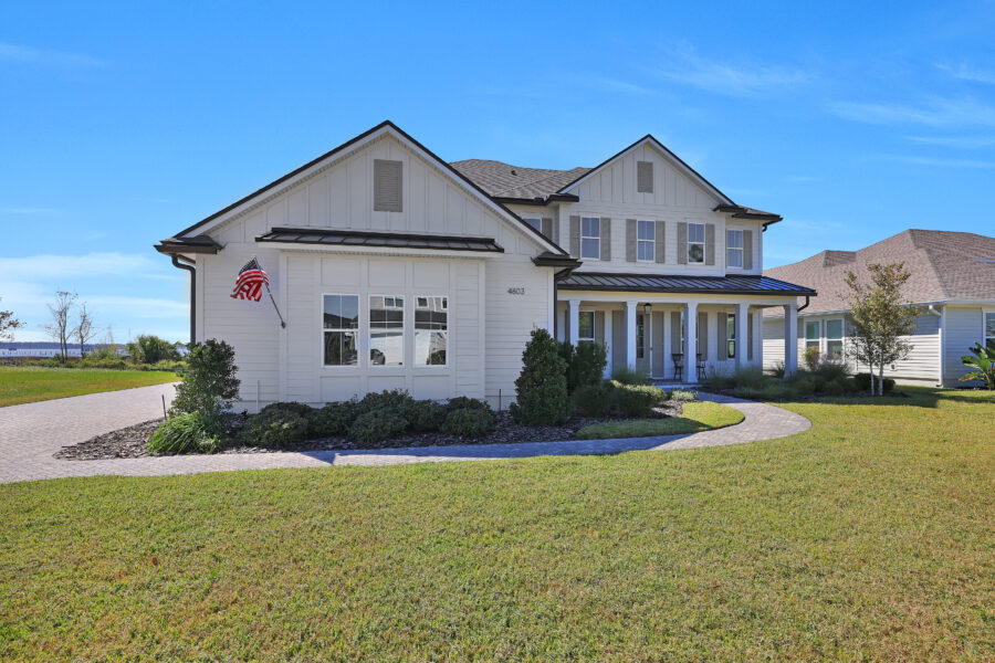 Bright white two-story American home with a spacious front porch, well-maintained lawn, and landscaped garden under a clear blue sky, showcasing modern farm-style architecture.