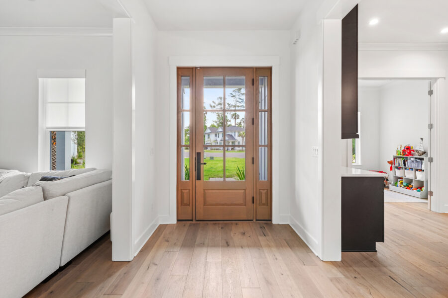 Bright modern home entryway with wooden front door and white interior walls, leading to living space and child's playroom with colorful toys.