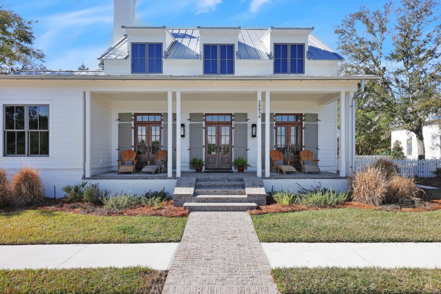 Modern white farmhouse with a spacious front porch featuring wooden chairs, planters, and brick steps, set against a bright blue sky with well-maintained landscaping and mature trees.