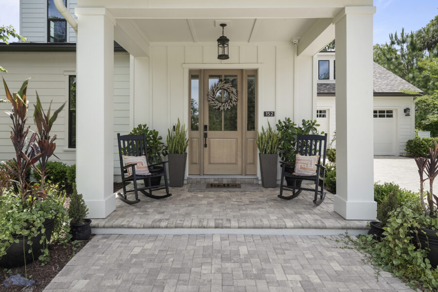 Cozy front porch with two black rocking chairs, lush green plants in tall planters, and a welcoming "hello" doormat, featuring white siding, a wooden door with a decorative wreath, and a covered entryway.