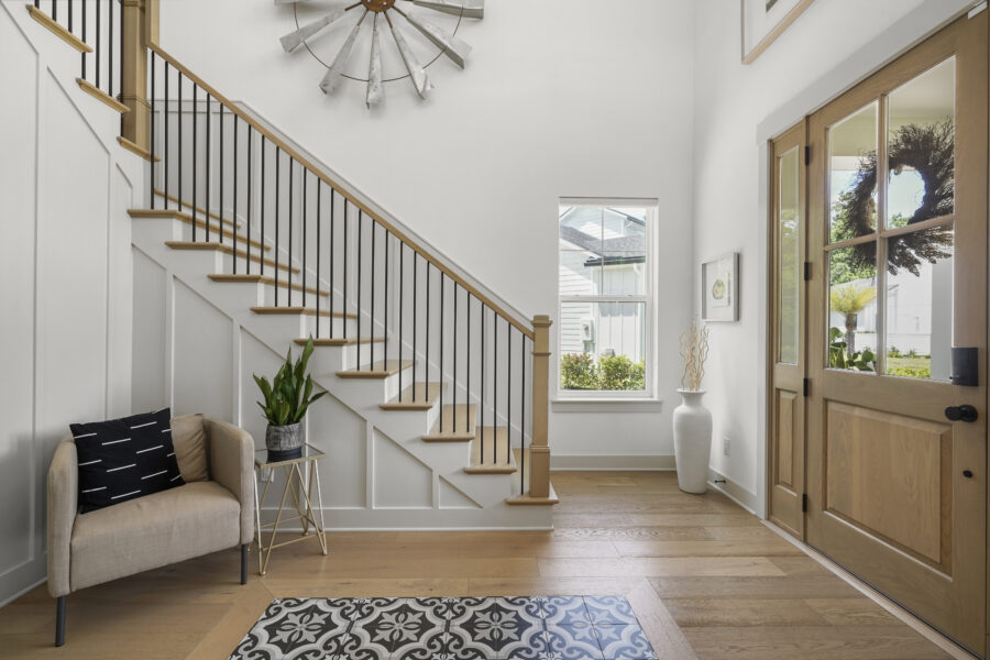 Bright entryway of a modern home featuring a wooden front door, a staircase with wooden steps and black metal railing, natural light from windows, and tasteful decor including a small sofa and potted plants.