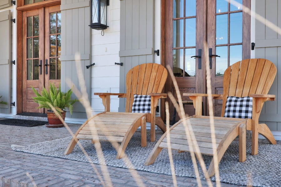 Adirondack wooden chairs with black and white checkered pillows on a porch with a textured rug and potted greenery.