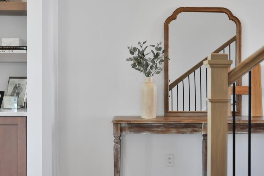 Elegant entryway with a rustic wooden console table, a tall cream-colored vase with greenery, and a large wooden-framed mirror reflecting a staircase with black iron balusters.