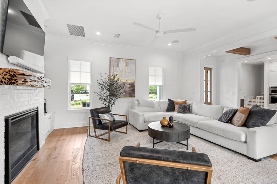 Modern living room with white walls, a cozy sectional sofa, black accent chairs, a fireplace with a white brick surround and wood mantel, and plenty of natural light from large windows.
