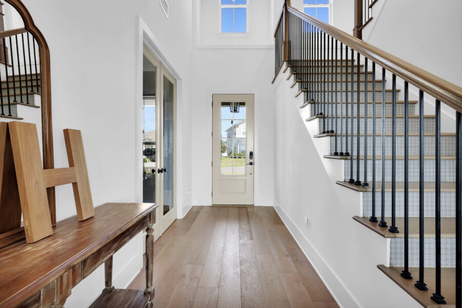 Bright interior view of a modern home's entryway featuring a staircase with wooden steps and black iron balusters, white walls, and a glass-paneled door leading outside, with ample natural light.