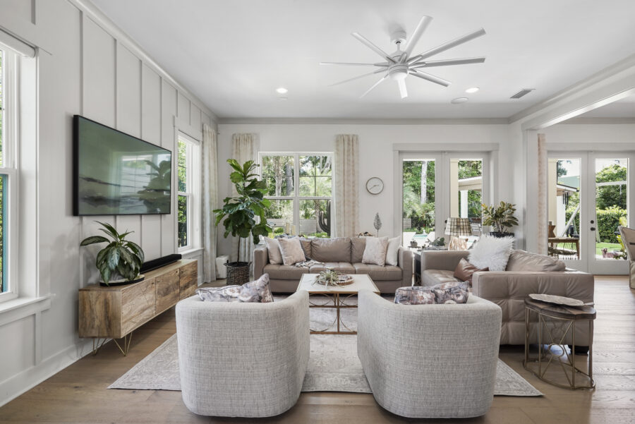 Modern living room with neutral-colored sofas, armchairs, a large flat-screen TV, and abundant natural light from multiple windows and glass doors, overlooking a lush backyard.