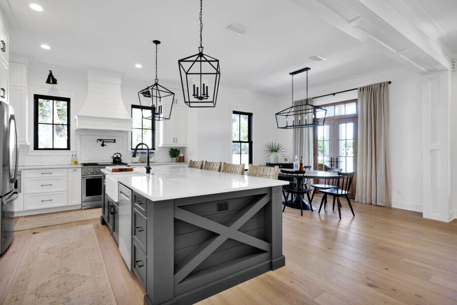 Modern white and black kitchen with farmhouse style accents, pendant lighting, and natural hardwood floors, featuring an island, stainless steel appliances, and large windows with curtains.