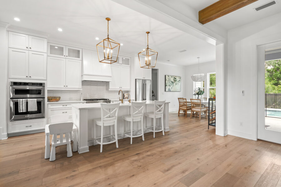 Modern white kitchen with stainless steel appliances, wooden flooring, and cozy breakfast bar, opening to a bright dining and living area with large windows and outdoor access.