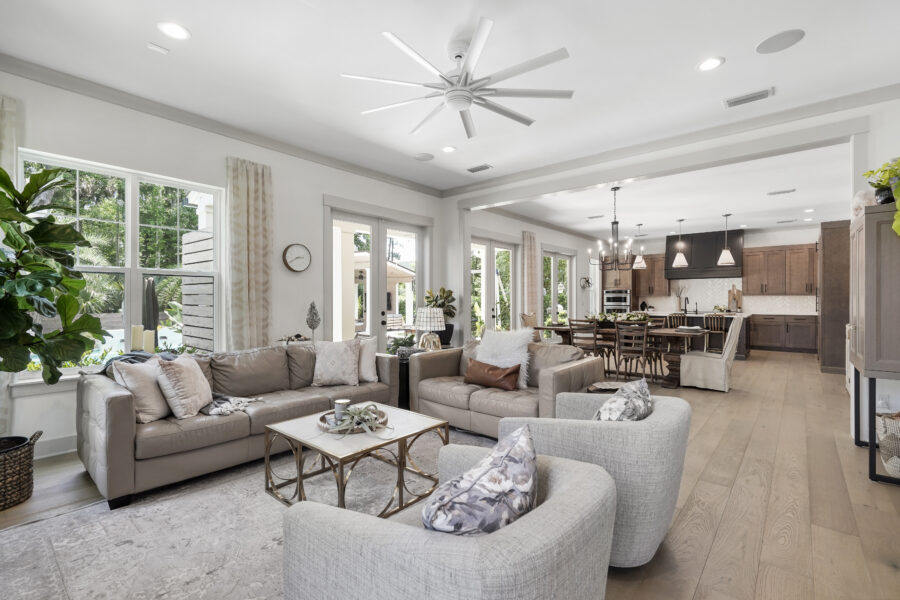 Spacious living room with large windows, neutral-toned sofas, a cozy armchair, and decor accents, seamlessly connected to an open kitchen and dining area with modern finishes and a view of the backyard.