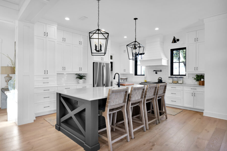 Modern white kitchen with a large island, black fixtures, pendant lighting, and natural light from windows, showcasing contemporary design and spacious layout.