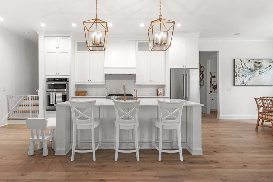 Modern white kitchen with an island, gold pendant lighting, and seating; spacious and bright interior with minimalistic decor and wooden flooring.