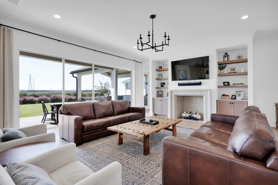 Spacious living room featuring leather sofas, a rustic wooden coffee table, built-in white shelves, a fireplace, and a large flat-screen TV, with expansive sliding glass doors leading to an outdoor patio with scenic views.