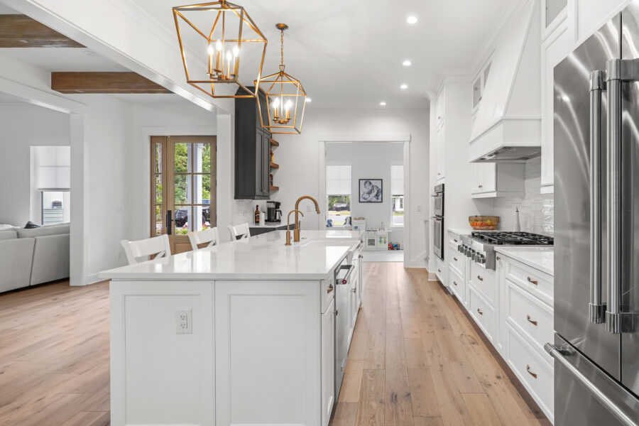 Contemporary white kitchen with island, gold fixtures, hardwood floors, and modern appliances, featuring open layout and stylish lighting.
