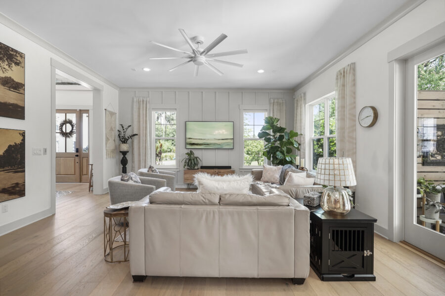 Bright and airy living room featuring large windows, white walls, a modern ceiling fan, and comfortable neutral-toned furniture with decorative plants and artwork.