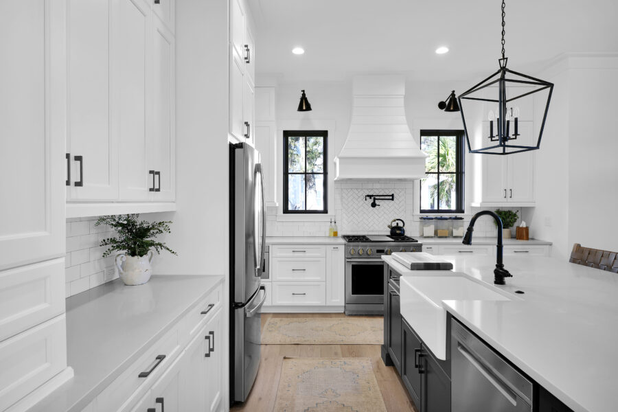 Modern white and black kitchen with farmhouse sink, stainless steel appliances, black hardware, and large windows providing natural light.