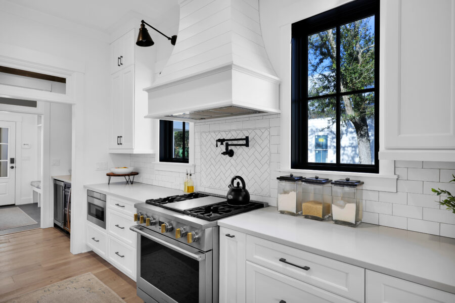 Bright modern kitchen with white cabinetry, black window frames, stainless steel stove, and decorative tile backsplash; natural light streams through large windows overlooking the outdoors.