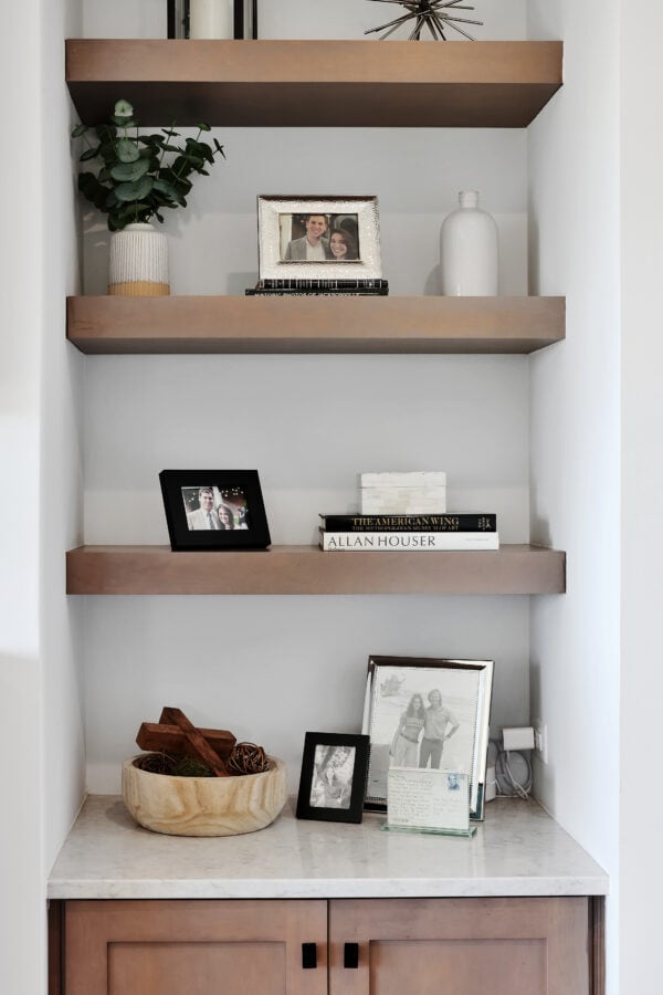 Photo of decorative shelving with framed family photos, books, and decorative items in a modern home interior alt text depicting wall shelves with framed family photos, books, and decorative objects in a contemporary home setting caption Stylish wall shelves with family photos, books, and decorative accents in a modern home interior image description The image showcases a built-in shelving unit with three wooden shelves holding framed family photos, books, and decorative items, set against a white wall. A marble-topped cabinet below completes the decor, blending modern style and personal touches in a cozy, elegant living space. relevance Highly relevant.