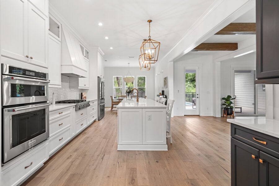 Modern white kitchen with a large island, stainless steel appliances, wooden flooring, and ample natural light from windows and glass doors. Elegant, bright, and spacious design.