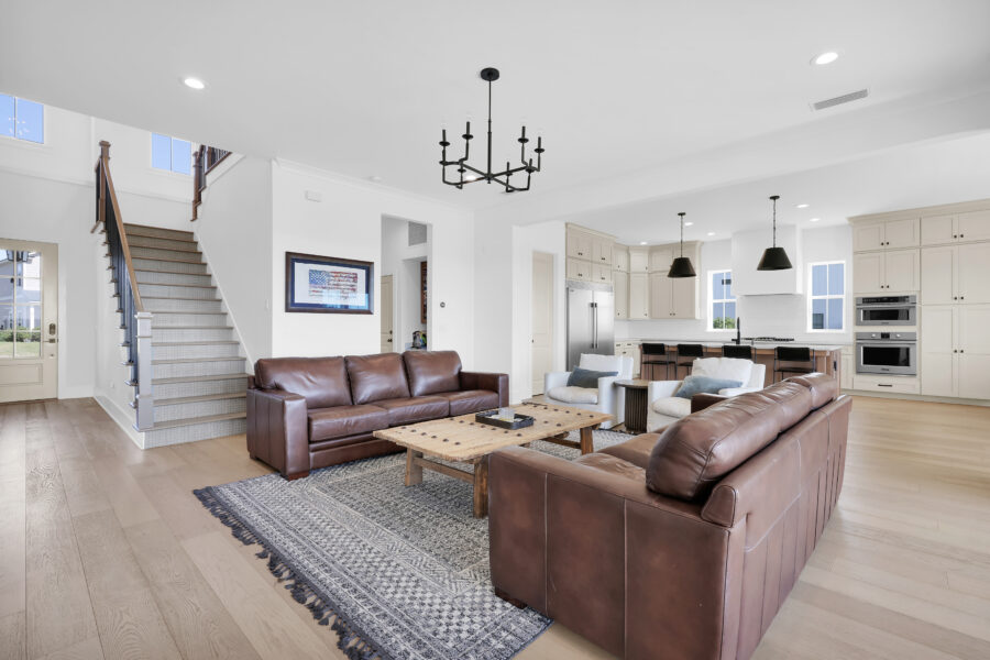 Spacious open-concept living room with leather sofas, a rustic wooden coffee table, and modern decor, flowing into a bright kitchen with white cabinetry and black pendant lights.