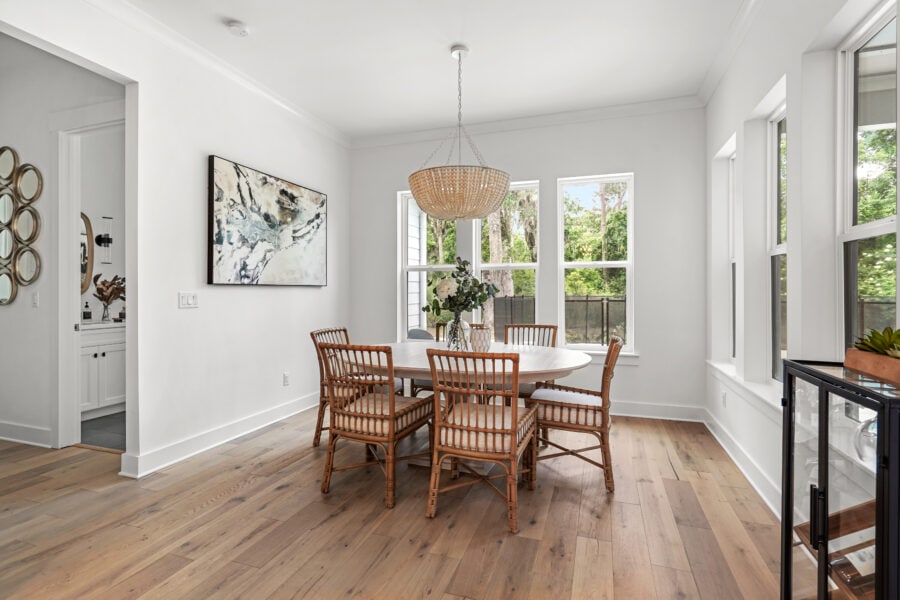 Bright dining room with white walls, large windows for natural light, wooden flooring, a round dining table with six wicker chairs, and a woven pendant light overhead.
