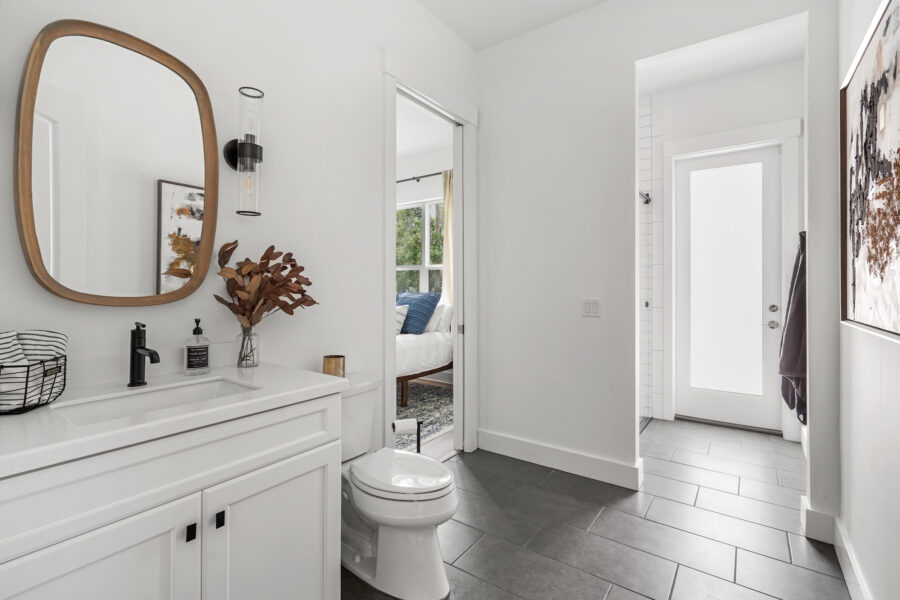 Modern white bathroom with a sleek vanity, black fixtures, and a decorative mirror, leading to a bedroom with natural light and stylish decor, showcasing contemporary home design.