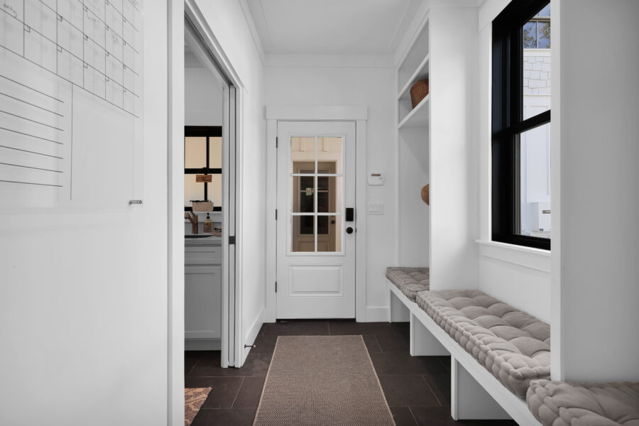 White entryway with built-in bench, black-framed window, and front door with glass panes; transitions into mudroom or foyer area in a modern home.
