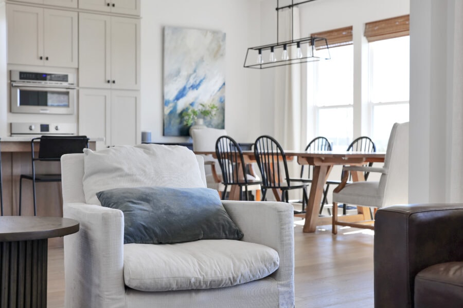 Neutral-toned living room with white sofa, blue pillow, open kitchen, dining area with wooden table and black chairs, large windows with blinds, and modern decor emphasizing natural light and spaciousness.