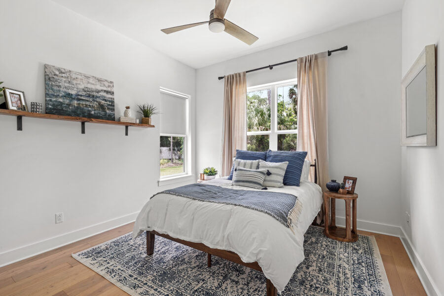 Bright bedroom with white walls, large windows with beige curtains, a wooden bed with white and blue bedding, and modern decor for a cozy, stylish home.