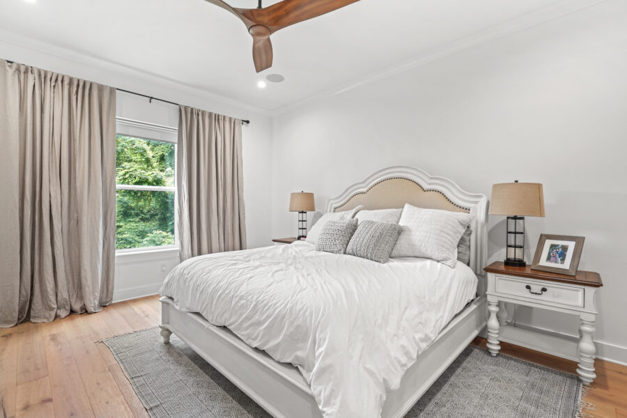 Spacious bedroom with a white bed, neutral pillows, and elegant curtains, creating a cozy and modern atmosphere. Natural light illuminates the room, highlighting the wooden floors and classic decor.