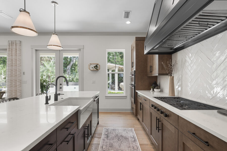 Modern kitchen with white countertops, wooden cabinets, a large farmhouse sink, black fixtures, and a stylish herringbone tile backsplash, illuminated by pendant lights and natural light from windows.