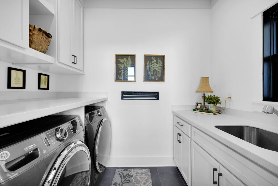 Modern laundry room with white cabinets, front-load washer and dryer, farmhouse-style decor, potted plant, and framed botanical art for a clean, functional space.
