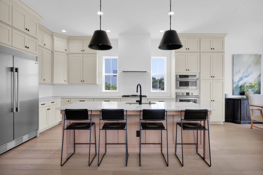 Modern white kitchen with large island, black pendant lights, built-in stainless steel appliances, and natural light from windows, showcasing stylish and functional home design.