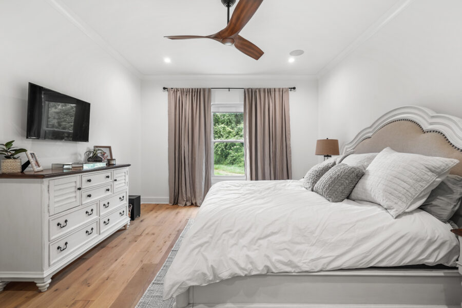 Modern master bedroom with white walls, cozy bedding, a decorative beige headboard, wooden flooring, a large window with beige curtains, and stylish furnishings including a white dresser and wall-mounted TV.