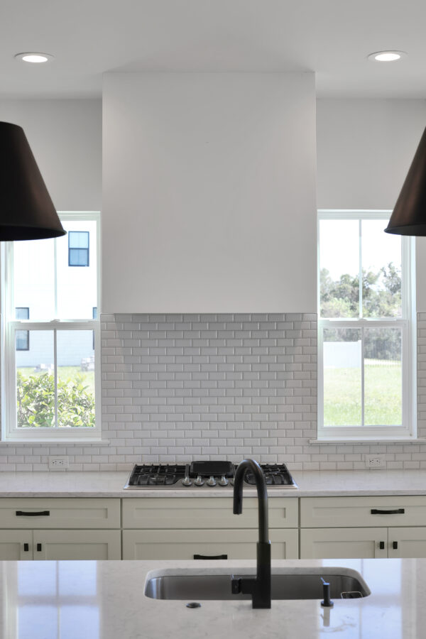 Modern kitchen with white subway tile backsplash, black fixtures, and large windows allowing natural light. The space features white cabinetry, a gas stove, and a clean, minimalist design.