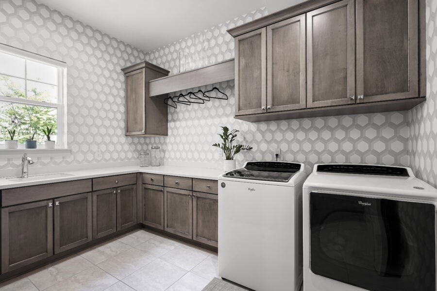 Modern laundry room with gray wooden cabinets, a washer and dryer, a window with potted plants, and white geometric wallpaper creating a bright, stylish space.