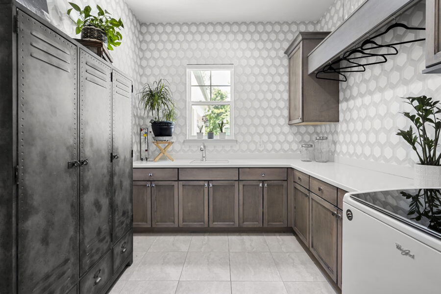 Modern kitchen with wood cabinetry, white countertops, decorative tile backsplash, and a small window bringing natural light. It features plants, glass jars, and appliances, blending functionality with stylish design.