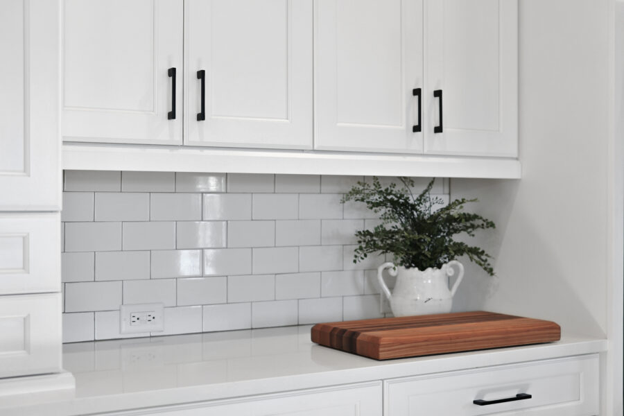 Modern white kitchen cabinetry with black handles, a white subway tile backsplash, a wooden cutting board, a wall outlet, and a decorative green plant in a white ceramic vase.