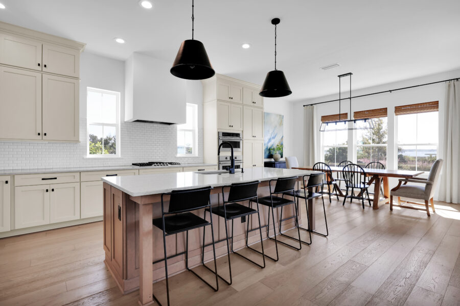 Modern open-concept kitchen with white cabinetry, black pendant lighting, and a large island with seating, seamlessly connected to a dining area with expansive windows overlooking scenic views.