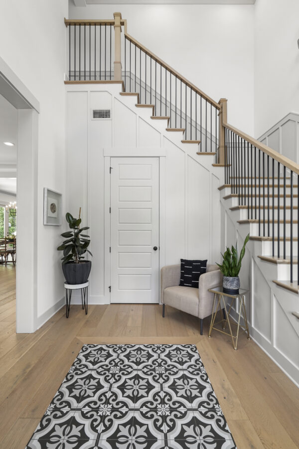 Staircase with black iron railings, wooden handrail, white paneled wall, potted plants, beige armchair, patterned rug, and light hardwood flooring in a modern home interior.