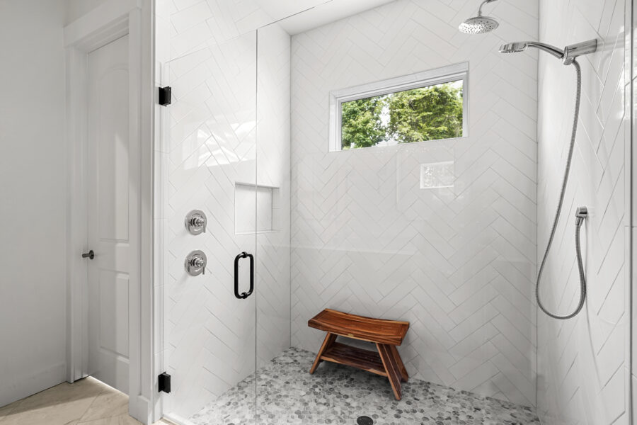 Modern walk-in shower with white herringbone tile walls, pebble floor, a built-in niche, a wooden bench, and a window for natural light, showcasing elegant bathroom design by John Merrill Homes.