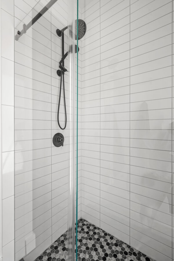 Shower with black fixtures and a glass partial door, surrounded by white tiled walls with black and white hexagonal tile flooring.