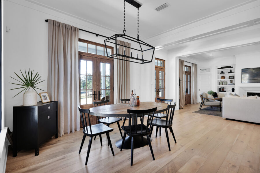 Modern dining area with a round wooden table, black chairs, large windows with wooden trim, and elegant curtains, bathed in natural light.