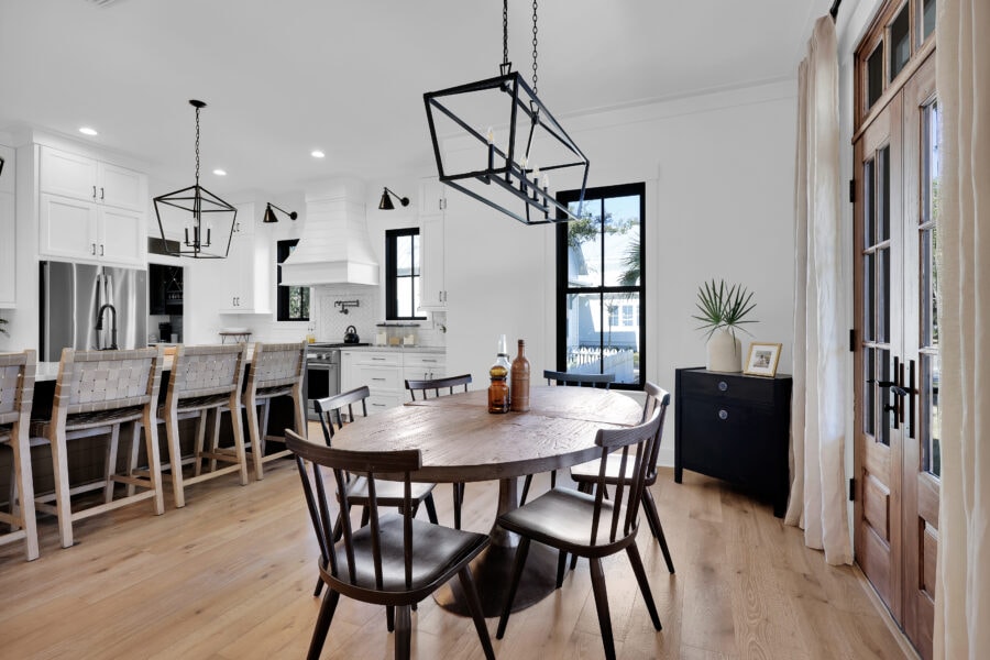 Modern farmhouse kitchen and dining area with white cabinetry, black accents, and natural wood furniture, featuring large windows and a contemporary light fixture.