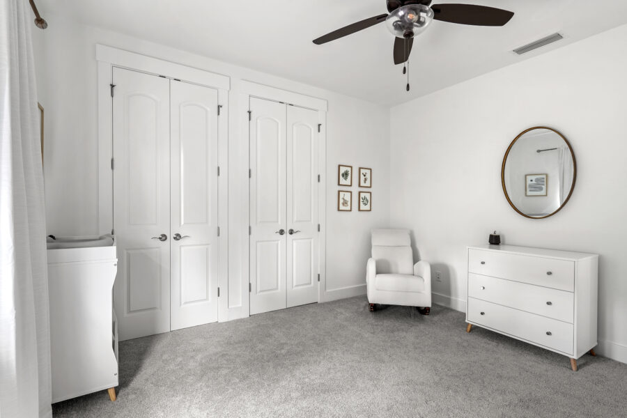 Custom white bedroom with closet doors, gray carpet, white dresser, armchair, and minimalist wall decor. Ceiling fan adds functionality and style to this bright, modern space.