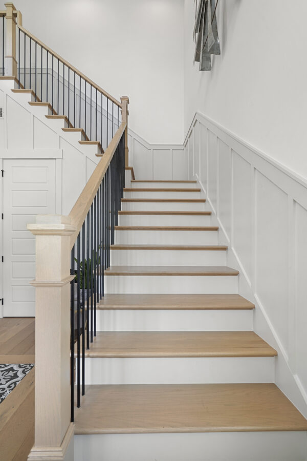 Wooden staircase with black metal balusters and white paneled walls, leading upstairs in a modern home.