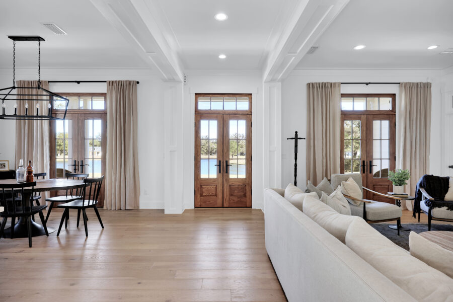 Bright open living and dining area with wooden double doors, large windows, neutral-colored sofa, light wood floors, and elegant decor, showcasing modern farmhouse interior design.